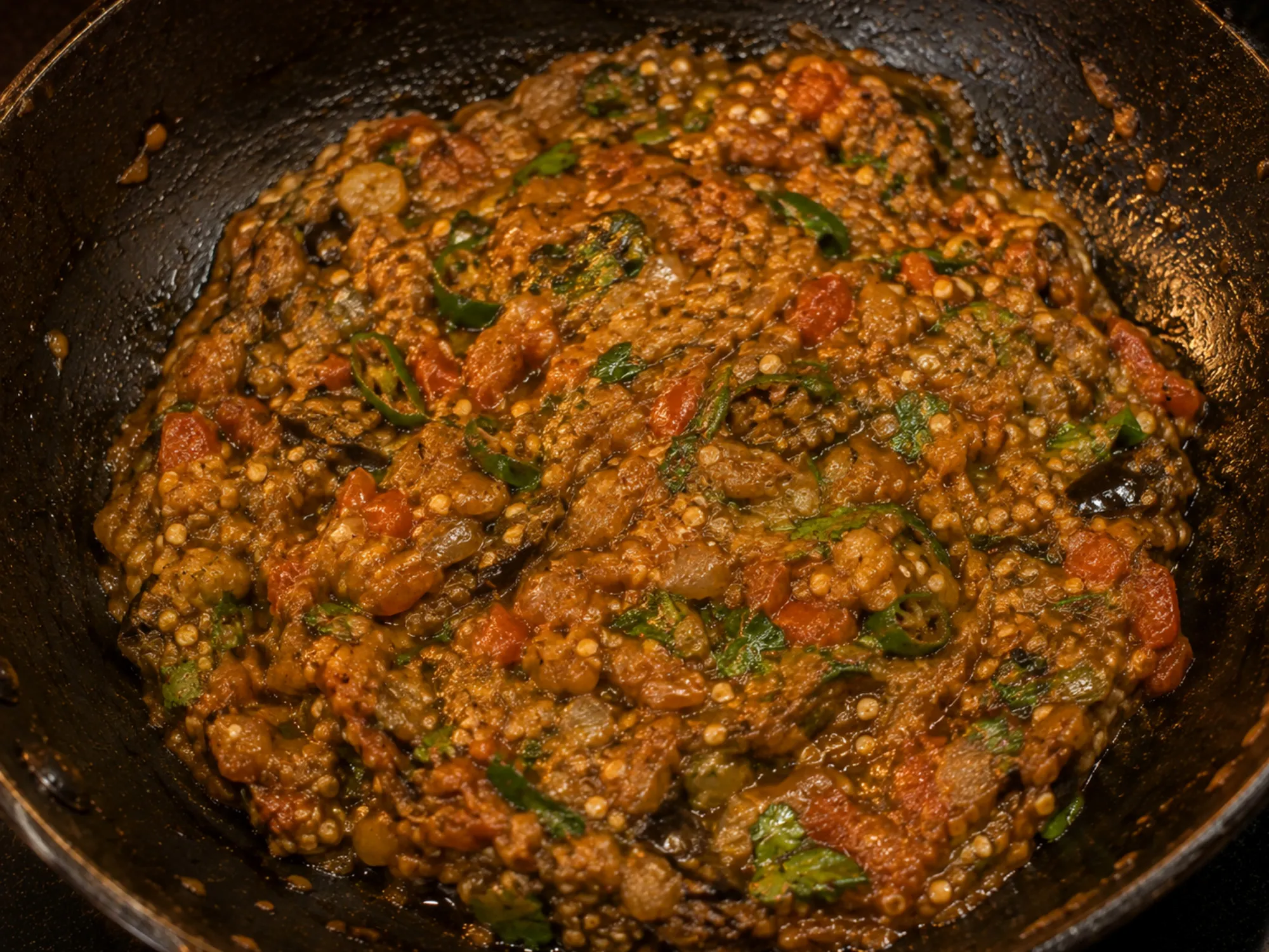 A dark, coarsely mashed brinjal dish in a kadai, flecked with tomato and green chilli, coriander on top