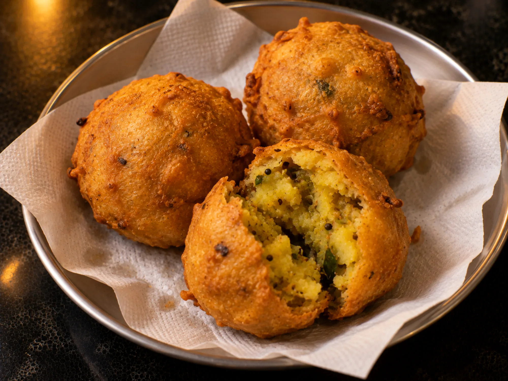 Round golden batata vada piled on a paper-lined steel plate, one split open to show the yellow potato filling