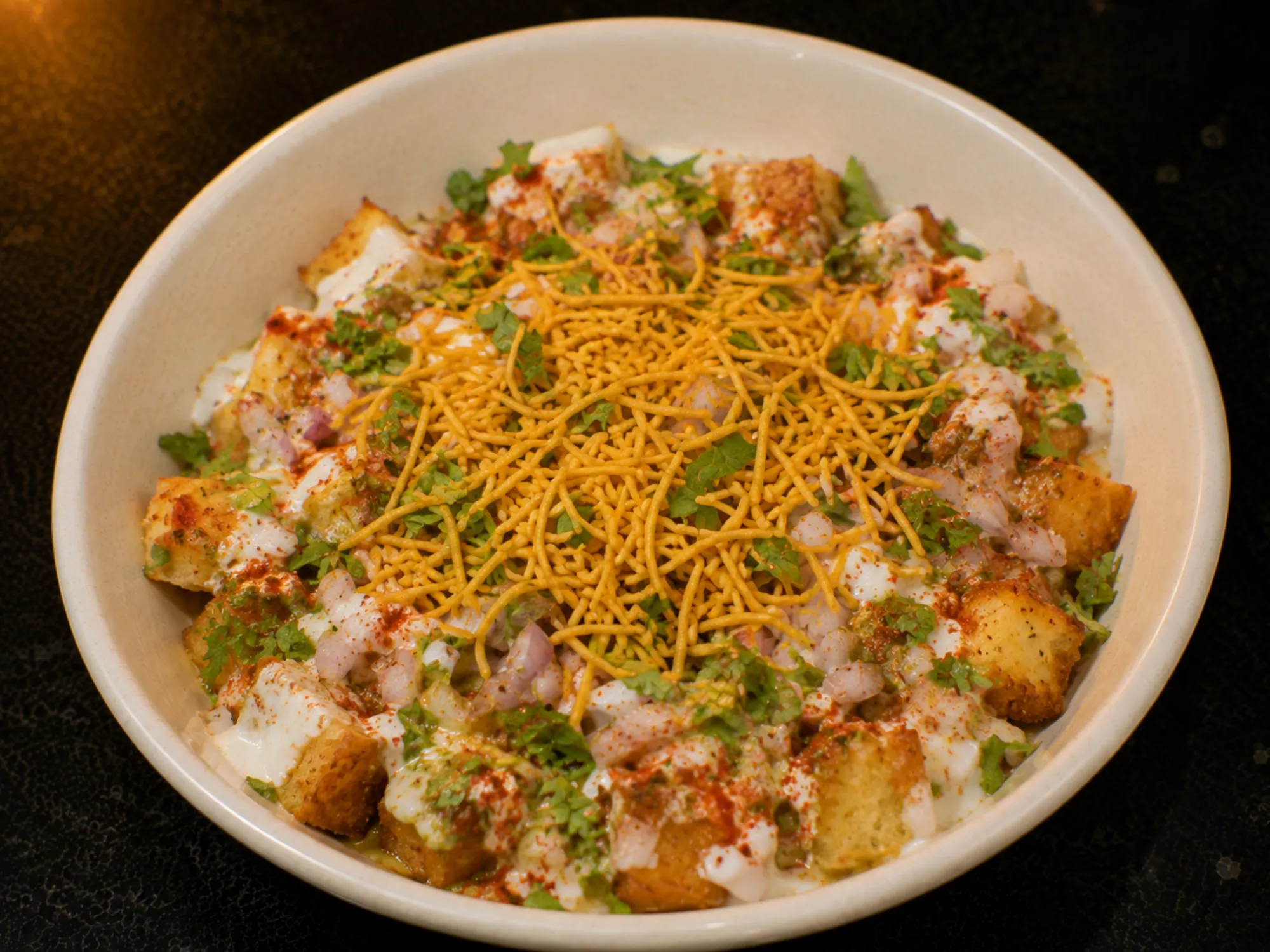 A bowl of golden bread cubes under white curd, red chilli powder, chopped onion, coriander and a pile of yellow sev