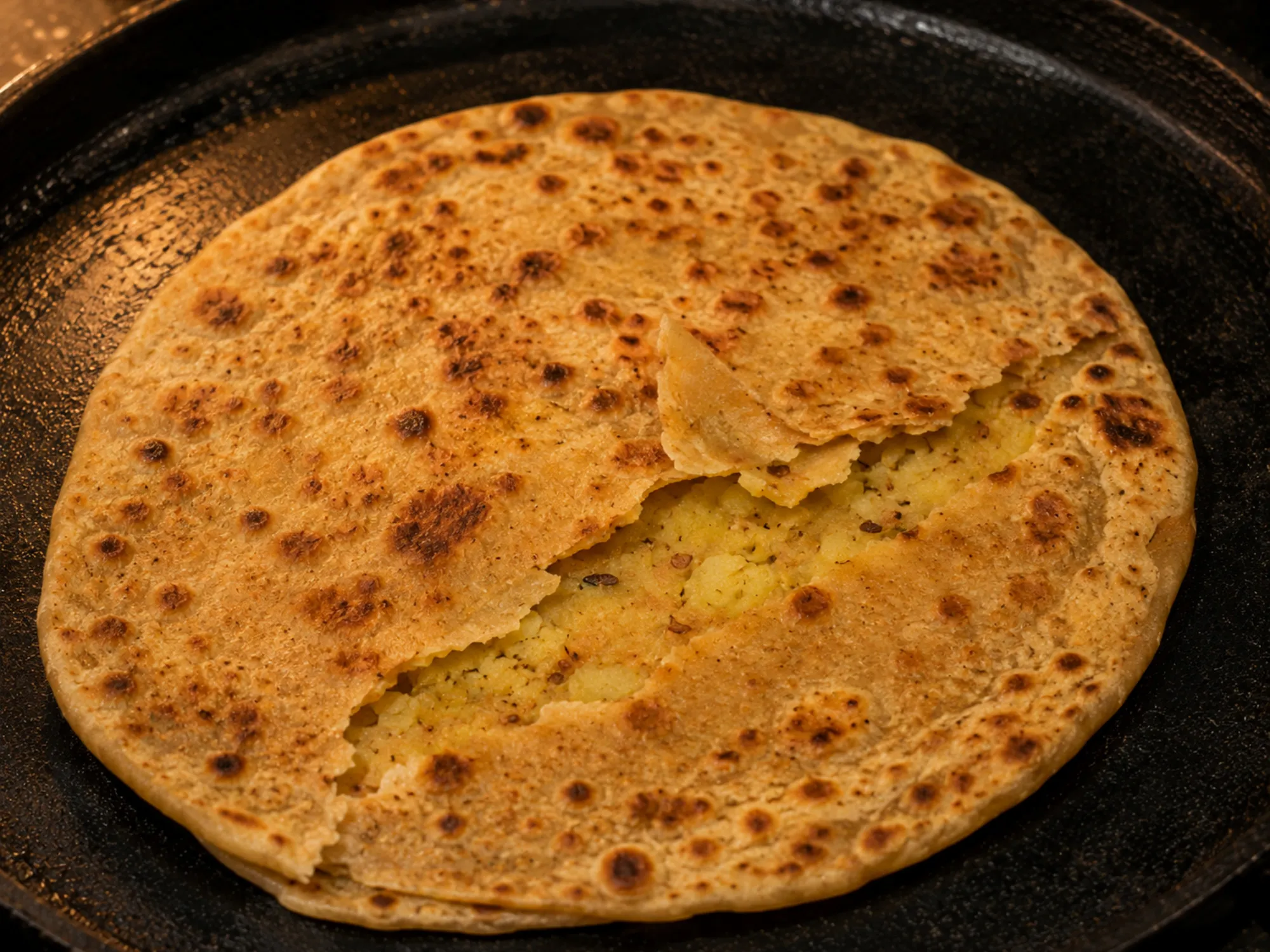 A golden-brown paratha on a tawa, blistered in patches, torn open to show a pale spiced potato filling