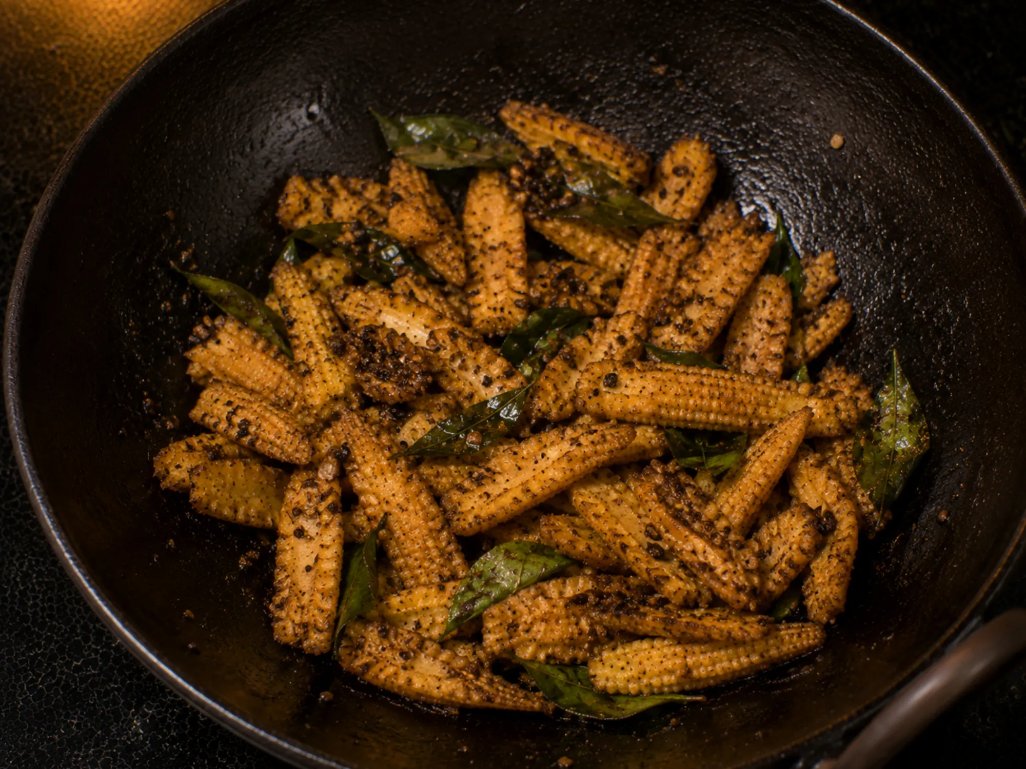 Browned halved baby corn coated in dark crushed pepper and curry leaves in a black kadai