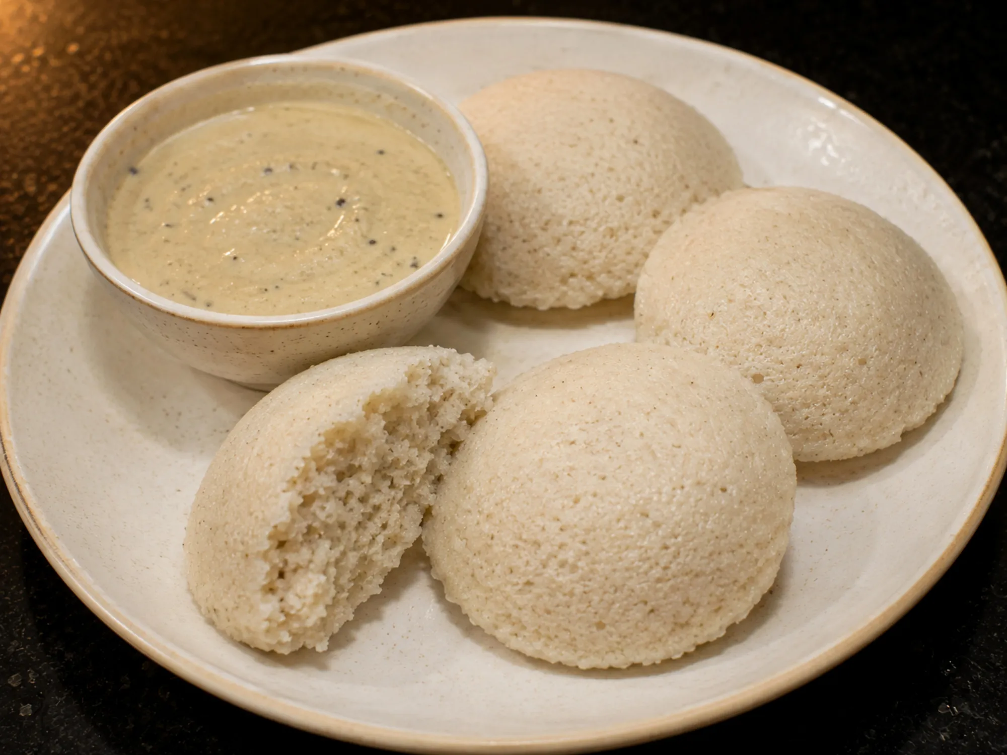 Four pale idlis on a plate beside a bowl of chutney, one broken to show the open crumb