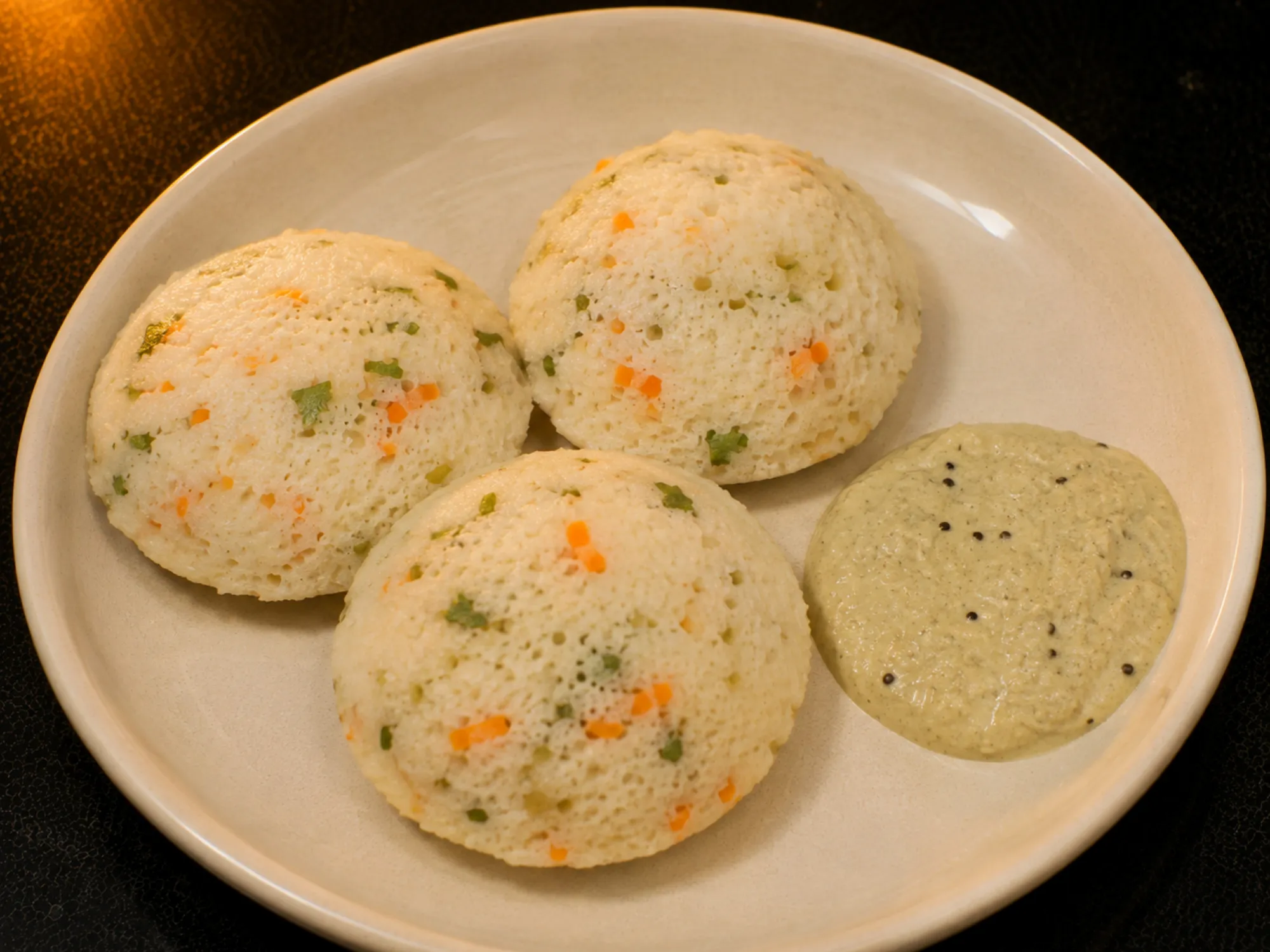Pale steamed idlis in a plate, flecked with orange carrot and green coriander, with chutney beside them