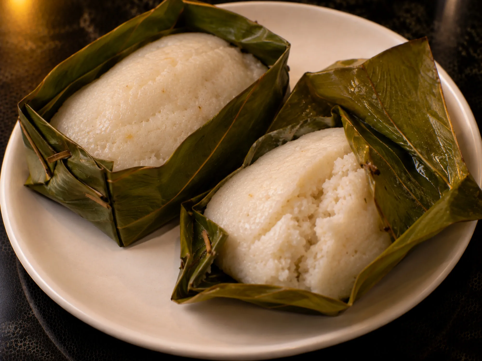 Steamed white kadubu being lifted out of a green jackfruit-leaf box, the leaf peeling away cleanly
