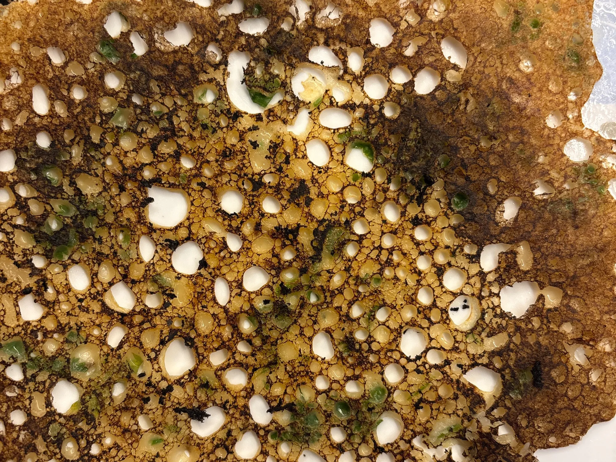 A thin lacy brown crepe on a tawa, full of irregular holes, flecked with cumin and coriander