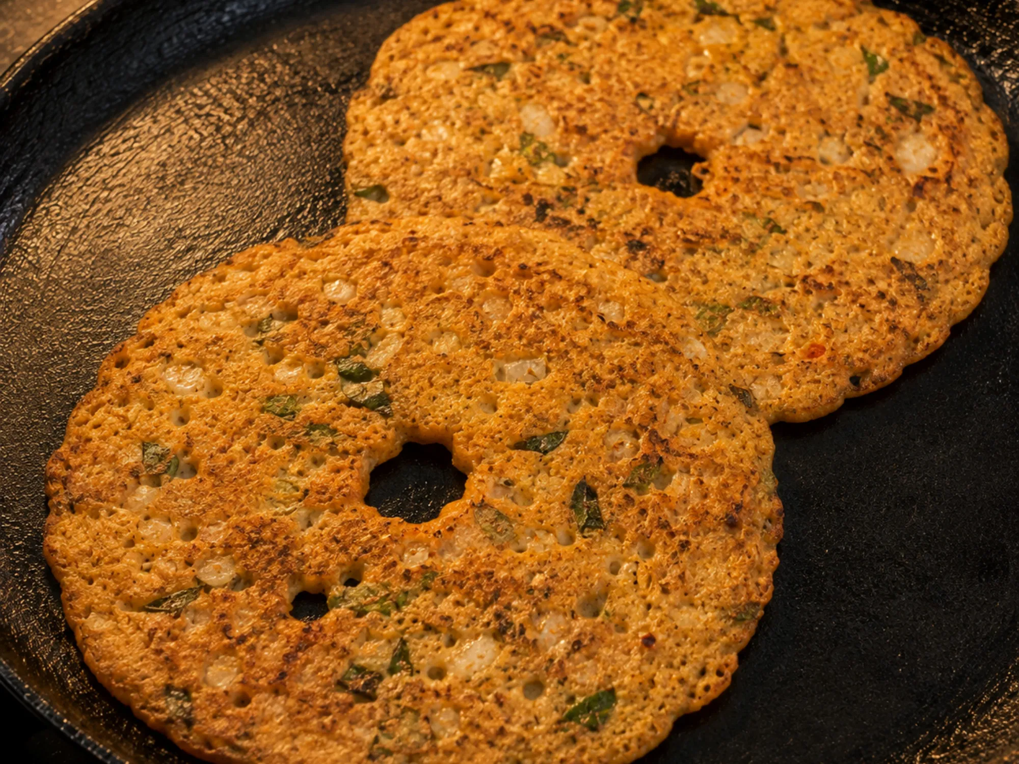 A thick brown adai on a tawa with a hole in the middle, flecked with onion and curry leaves