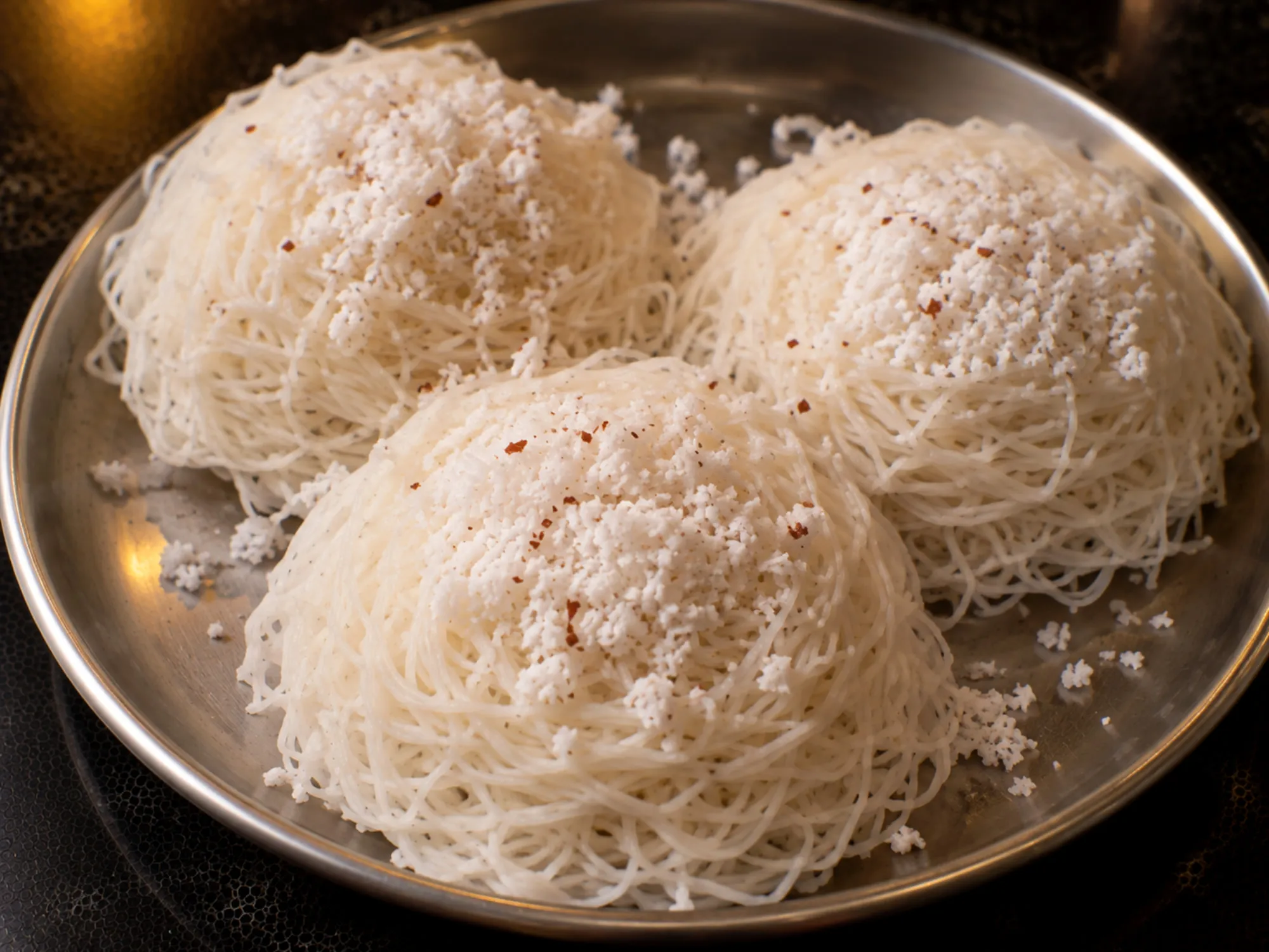 A stack of white idiyappam nests topped with grated coconut on a steel plate