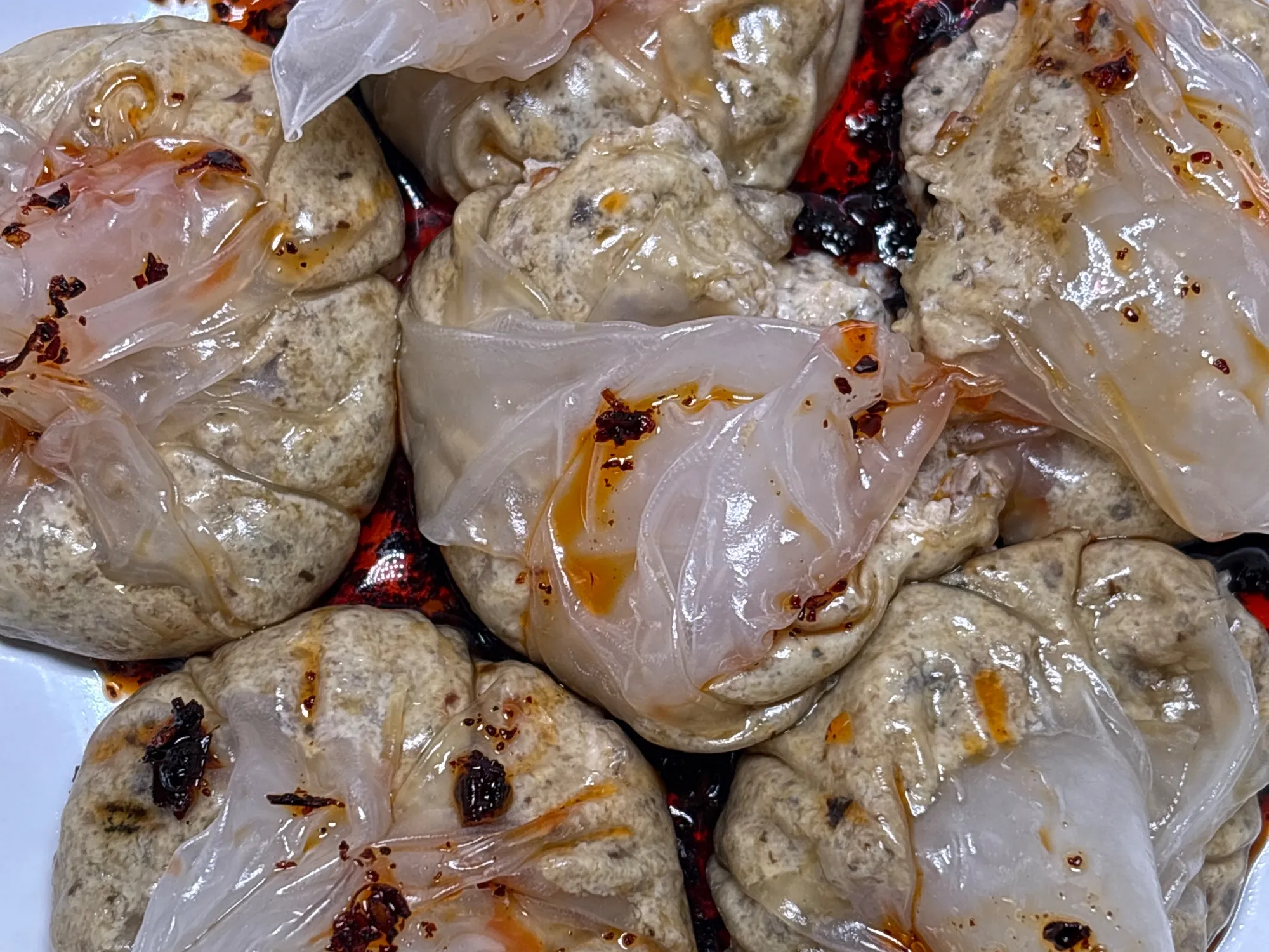 Four pleated steamed dumplings with translucent wrappers sitting in a pool of dark red chilli oil in a black bowl